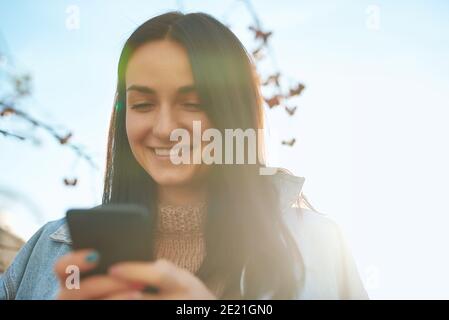 Primo piano di una giovane femmina con un sorriso brillante comporre il numero sul suo telefono cellulare su uno sfondo sfocato alla luce del sole Foto Stock