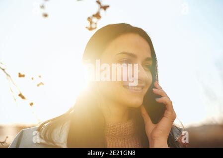Primo piano ritratto di una giovane femmina sorridente che la tiene smartphone vicino all'orecchio all'aperto in una calda serata autunnale Foto Stock