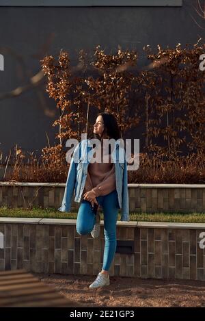 Bella giovane donna in denim vestiti in piedi in posizione rilassante in una piazza d'autunno, crogiolandosi in luce solare calda Foto Stock