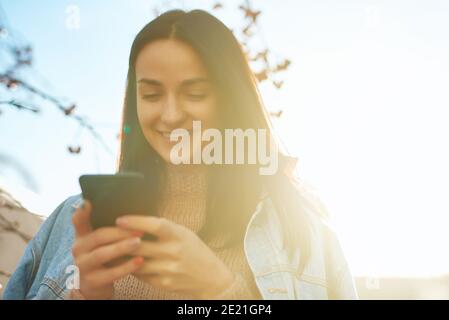 Allegra giovane donna sorridente che comunica via internet utilizzando un cellulare all'aperto in una bella giornata di sole in autunno Foto Stock