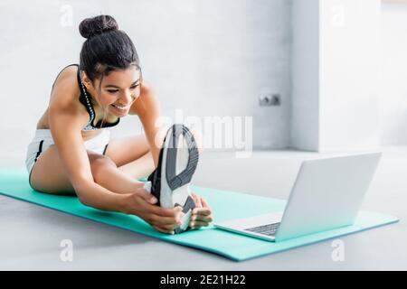 allegra donna afroamericana che stretching mentre si siede sul tappetino fitness vicino al computer portatile Foto Stock