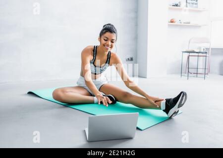 donna afroamericana sorridente in abbigliamento sportivo seduto sul tappetino fitness vicino al computer portatile a casa Foto Stock
