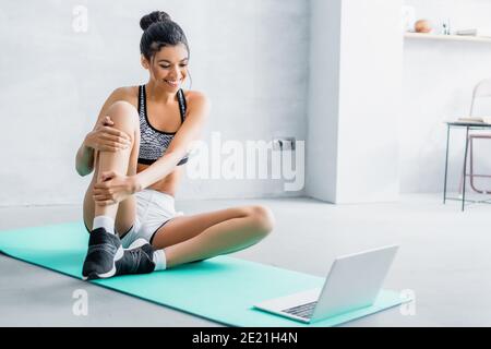 giovane afroamericana in sportswear sorridente mentre si siede sopra tappetino fitness vicino al computer portatile Foto Stock