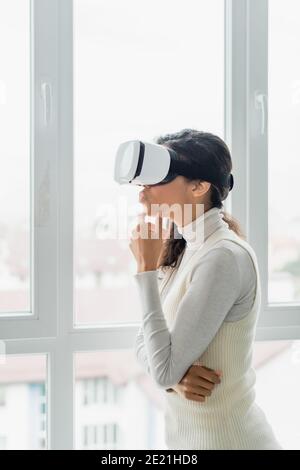 donna afro-americana con visore vr che guarda attraverso la finestra a casa Foto Stock