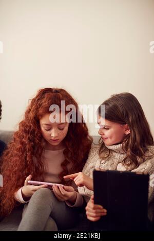 Gruppo di ragazze con amici seduti sul divano a casa Riproduzione su tablet digitale e telefoni cellulari Foto Stock