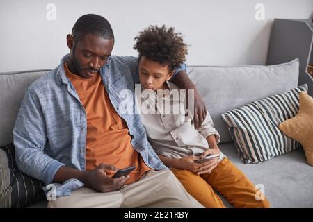 Uomo maturo moderno che indossa abiti casual seduto su divano con suo figlio teenager gli mostra le foto sullo smartphone Foto Stock