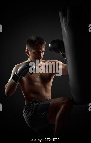 pugile muscolare in guanti di pugilato neri pugilato in sacchetto di boxe su sfondo scuro Foto Stock