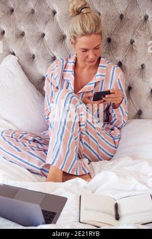 Donna d'affari in Pajamas seduta sul letto con laptop e cellulare Telefono funzionante da casa in blocco Foto Stock