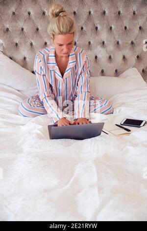 Donna d'affari in Pajamas seduta sul letto con il laptop che lavora da Casa durante il blocco pandemico Foto Stock