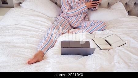 Primo piano di Businesswoman in Pajamas seduta sul letto con Computer portatile e telefono cellulare che funzionano da casa Foto Stock