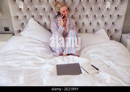 Donna d'affari in Pajamas seduta sul letto che fa la chiamata sul cellulare Telefono funzionante da casa Foto Stock