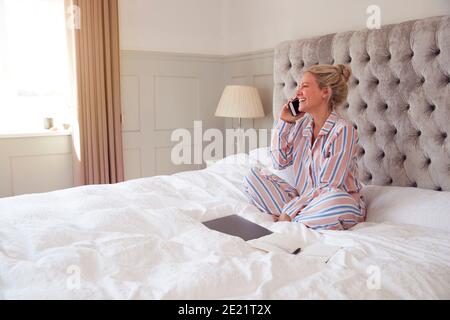 Donna d'affari in Pajamas seduta sul letto che fa la chiamata sul cellulare Telefono funzionante da casa Foto Stock