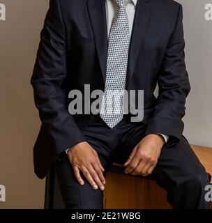 Uomo irriconoscibile dall'aspetto bello che indossa un abito nero e una cravatta, seduto su una sedia di legno e in posa Foto Stock