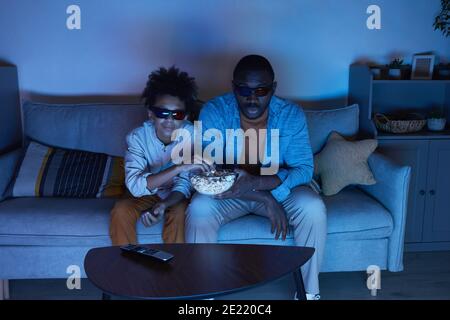 Alto angolo di scatto di afroamericano uomo e il suo teen Figlio seduto sul divano in soggiorno guardando film 3D e mangiare popcorn di sera Foto Stock