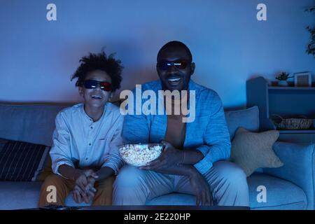 Ritratto dell'uomo afroamericano e del suo figlio teen seduto Insieme sul divano in salotto ridendo mentre si guarda il 3D film e mangiare popcorn Foto Stock