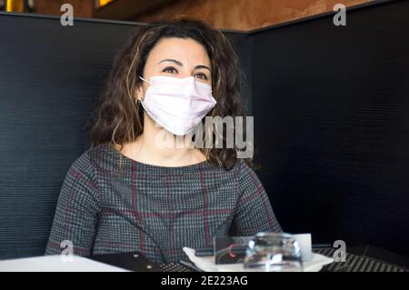 Ritratto di giovane donna che indossa una maschera medica, mentre si siede al tavolo nel ristorante Foto Stock