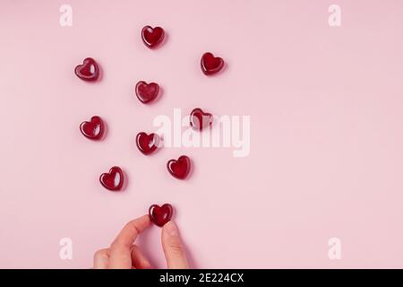 Mano che tiene una delle caramelle sparse di gelatina rossa del cuore forma su sfondo rosa Foto Stock