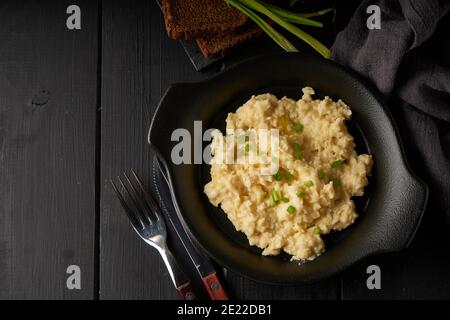 Uova strapazzate con cipolla verde servite in piatto scuro con pane sul tavolo nero Foto Stock