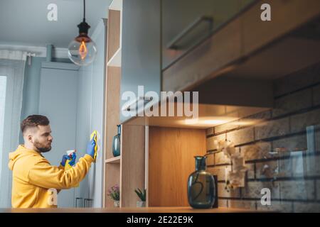 Vista laterale di un uomo caucasico con pulizia della barba la sua casa disinfettandola con spray indossando guanti Foto Stock