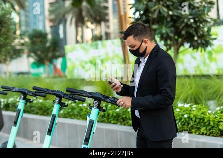 Scooter elettrico e telefono cellulare. Uomo d'affari che utilizza il servizio di noleggio auto con smartphone in città urbana strada e parco. Giovane ragazzo millenario che usa Foto Stock