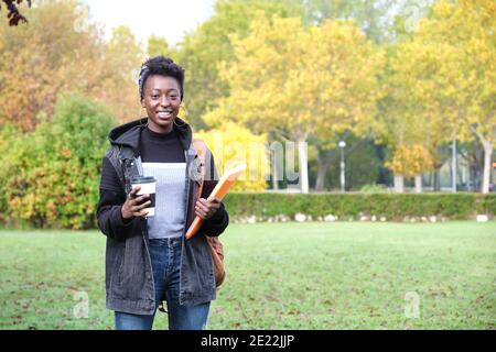 Studentessa africana universitaria con zaino, piegatrice e una tazza di caffè all'esterno del campus. Concetto di vita dell'università. Foto Stock