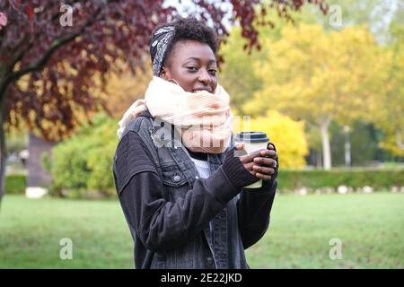 Bella donna africana che indossa una sciarpa e tiene una tazza di caffè in un parco. Concetti di autunno, inverno e freddo. Foto Stock