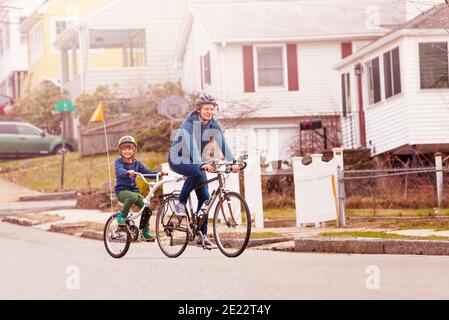 Ritratto in bici per tutta la famiglia con un piccolo ragazzo felice la bicicletta tandem di traino è collegata al padre sul lato urbano della strada visualizza Foto Stock