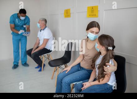 Madre e figlia indossano maschere per il viso, seduti in fila nella sala d'attesa dell'ospedale prima della vaccinazione con il coronavirus Foto Stock
