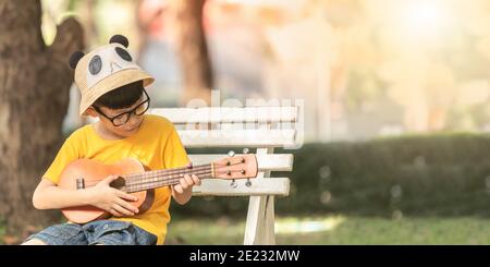 Un bambino asiatico indossare occhiali è felice di giocare il ukulele. Bambino asiatico sta cercando di giocare l'ukulele con un momento completamente felice. Foto Stock