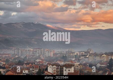 Misty, fumoso paesaggio urbano di Pirot durante il tramonto con impressionante, picco roccioso schiarita dalla luce dorata sullo sfondo e un cielo nuvoloso Foto Stock