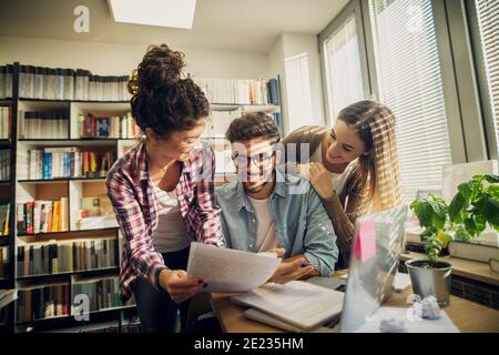 Due ragazze allietate allegre che aiutano il loro amico a passare exam.sitting in una biblioteca luminosa. Foto Stock