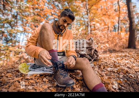 Hiker che legano la racchina mentre si siede a terra. Accanto a lui zaino. Boschi in autunno esterno, alberi e foglie cadute tutto intorno. Foto Stock