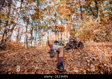 Escursionista stanco seduto a terra. Accanto a lui zaino. Foresta in autunno esterno. Foto Stock