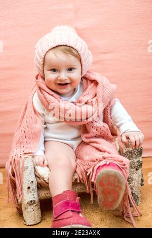 Primo piano viso bambino in snood. Cute baby girl con gli occhi blu in un cappello di maglia rosa e sciarpa su sfondo rosa.Cosy caldo inverno o primavera Foto Stock