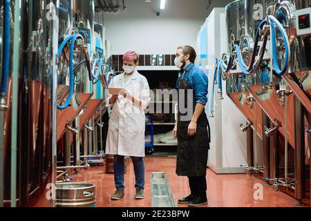 Giovane birreria femminile esperta in biancatura e maschera protettiva in ingresso dati in tavoletta mentre il produttore di birra maschile si trova vicino all'interno pianta Foto Stock