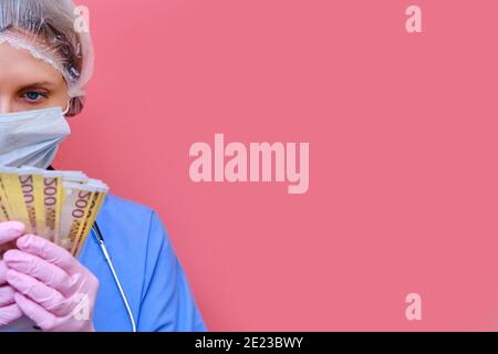 Un medico mantiene uno stipendio in euro, copia spazio per il testo. Un'infermiera con denaro per il lavoro e una maschera protettiva su sfondo rosa. Foto Stock