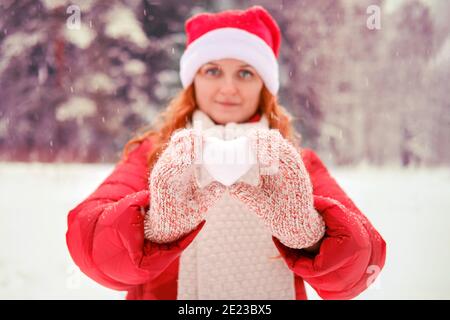 Donna nel cappello di natale con palla di neve a forma di cuore, fuoco selettivo sulle mani. Donna Redhead a Natale e Capodanno cammina all'aperto in un cappello rosso A. Foto Stock
