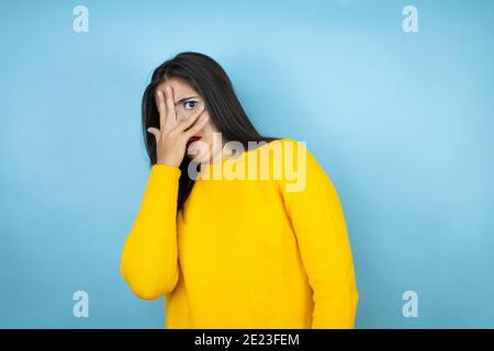 Giovane bella donna che indossa un maglione giallo su uno sfondo blu isolato sbirciando in shock coprendo viso e occhi con la mano, guardando attraverso le dita Foto Stock