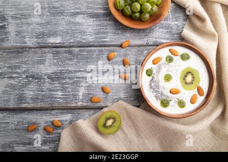 Yogurt con kiwi, uva spina, chia e mandorle in ciotola di legno su fondo grigio di legno e tessuto di lino. Vista dall'alto, piatto, spazio copia. Foto Stock