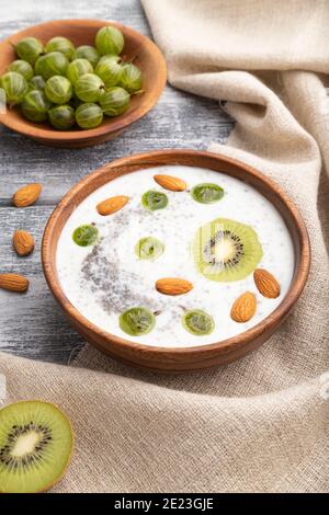 Yogurt con kiwi, uva spina, chia e mandorle in ciotola di legno su fondo grigio di legno e tessuto di lino. Vista laterale, primo piano. Foto Stock