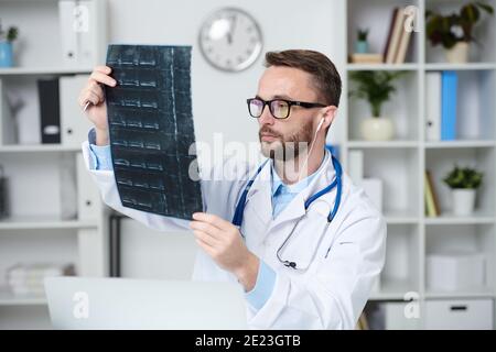 Giovane medico di sesso maschile serio in whitecoat, occhiali e auricolari seduti di fronte al computer portatile e guardando l'immagine radiografica del paziente durante la consultazione Foto Stock