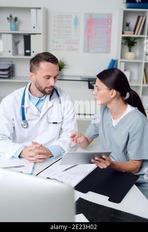 Due medici giovani e fiduciosi in uniforme seduti alla scrivania davanti al monitor del computer, discutono di documenti medici e consulenze Foto Stock