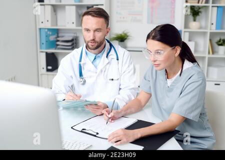 Due giovani medici in uniforme seduti davanti alla scrivania di monitor di computer e consulenza con colleghi più esperti in chat video Foto Stock