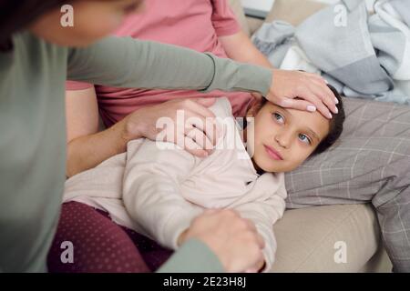 Mano di giovane donna attenta che si prende cura del suo malato adorabile figlia giacente sul divano sul padre si inginocchia mentre si tocca la sua fronte a casa Foto Stock