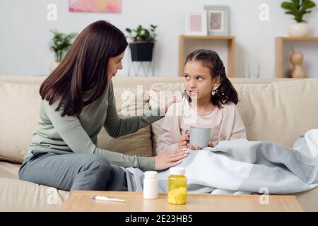 Giovane donna attenta che si prende cura della sua ammalata adorabile figlia seduto sul divano in soggiorno mentre si tocca la spalla e. dare la sua bevanda calda Foto Stock