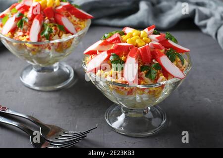 Due porzioni di insalata con bastoncini di granchio, uova e mais in coppe di vetro trasparenti su sfondo scuro. Primo piano Foto Stock