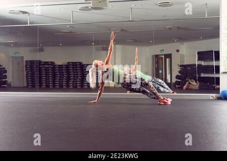 Montare l'atleta femminile in activewear facendo esercizio laterale tavola contro specchio in palestra Foto Stock