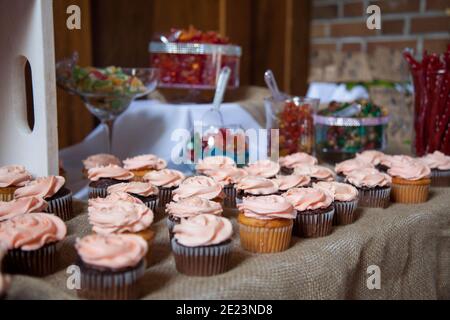 Vaniglia ghiacciata rosa e cupcake di cioccolato sedersi su un panno da tavolo di burlap con altre caramelle sullo sfondo come tavolo da dessert per un matrimonio. Foto Stock