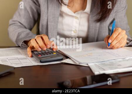 Una donna d'affari che indossa un abito formale sta lavorando in un ambiente d'ufficio. Sta calcolando i redditi e le spese usando le dichiarazioni, le polizze di paga, le ricevute. S Foto Stock