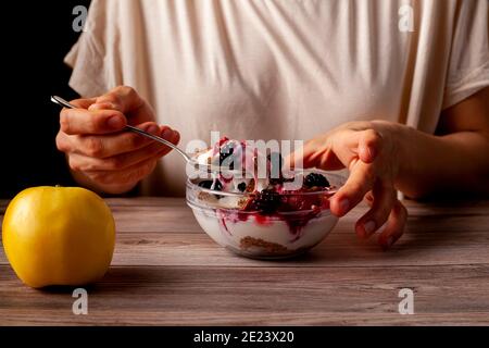 La donna sta mangiando una ciotola fresca fatta in casa di vetro di parfait cremoso di yogurt con bacche, muesli e semi come spuntino di notte. La foto mostra la mano che tiene a s. Foto Stock